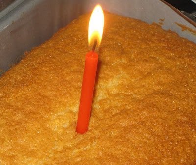 A single lit orange candle stands in the center of a rectangular, golden-brown cake in a baking tray.