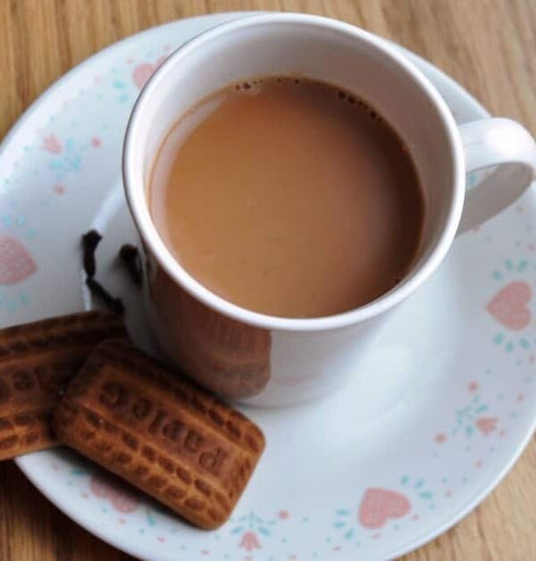 A cup of Indian Ginger Tea sits on a white, heart-patterned saucer with two rectangular cookies beside it.