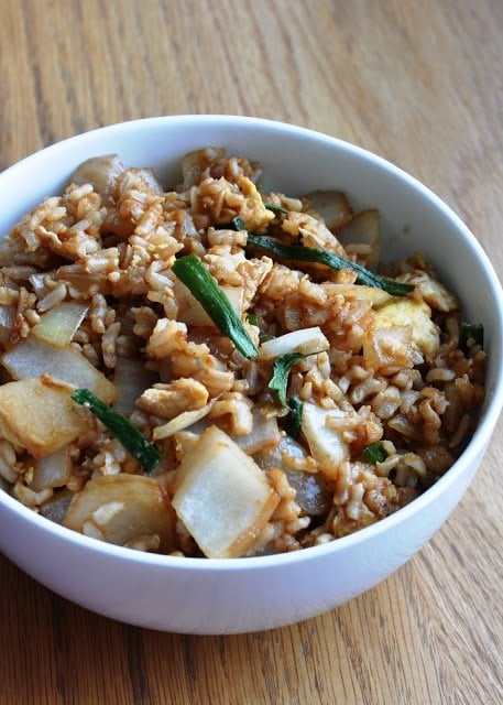 A bowl of quick and easy egg fried rice with onions, green onions, and scrambled eggs rests invitingly on a wooden table.