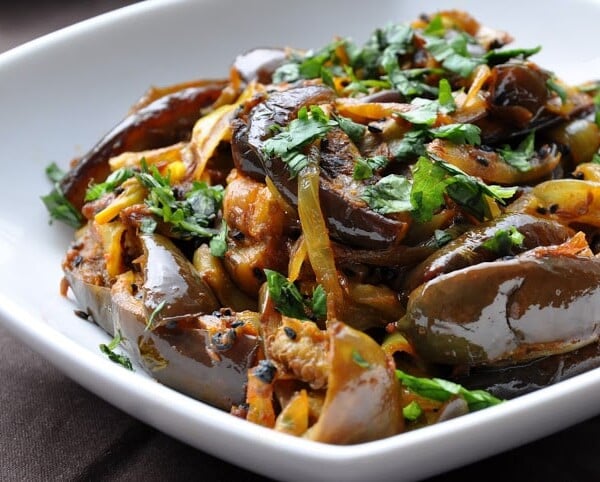 A dish of cooked eggplant slices mixed with onions and topped with chopped cilantro served in a white bowl.