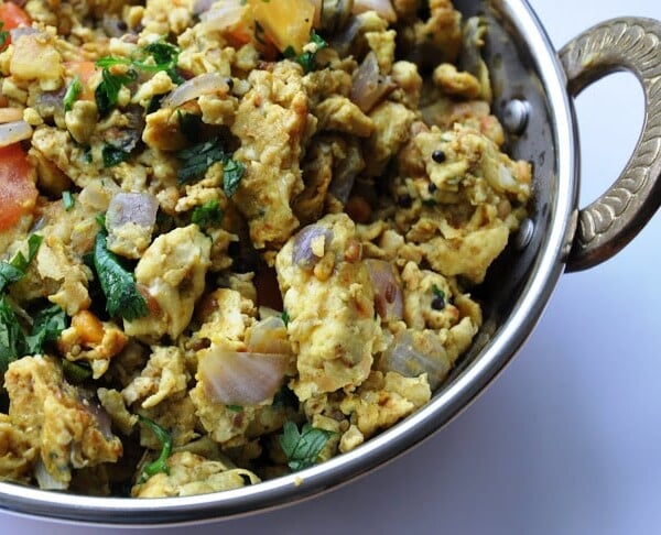 A dish of scrambled eggs with chopped onions, tomatoes, and herbs, served in a metal bowl with handles.