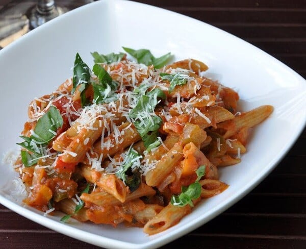 A bowl of penne pasta with tomato sauce, garnished with fresh basil and grated cheese, on a wooden table.