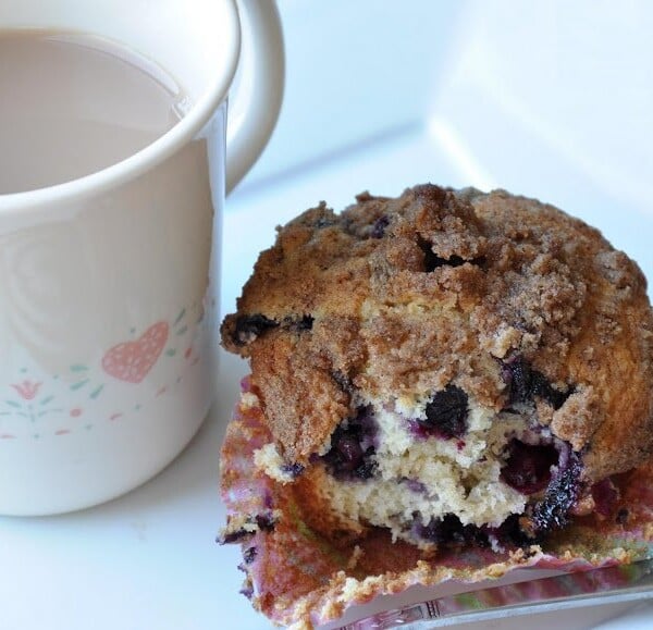 A partially eaten blueberry muffin with a crumb topping sits on a table next to a white mug filled with a light-colored beverage.