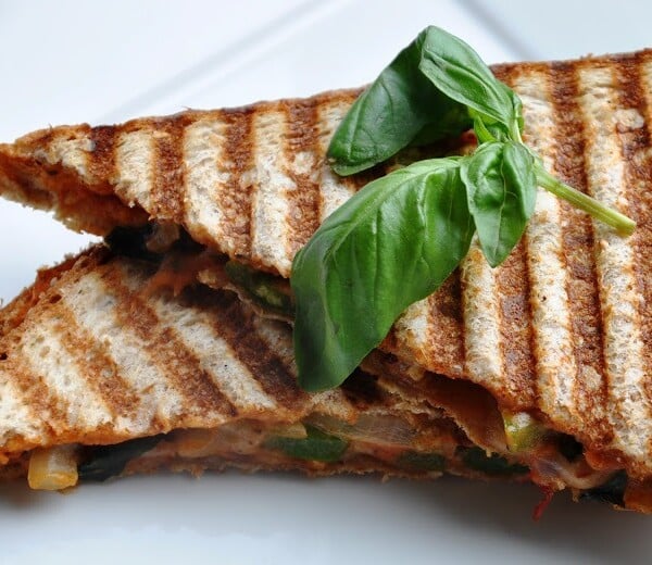 A grilled panini sandwich with visible grill marks, filled with vegetables, topped with fresh basil leaves on a white plate.