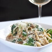 A bowl of Veggie Scampi pasta with broccoli and grated cheese, served on a white plate, with a glass of white wine in the background.