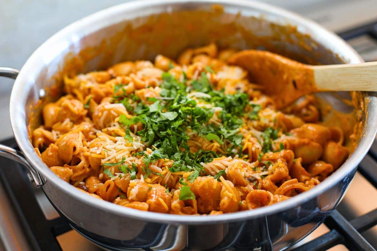 A skillet filled with Black Bean Enchilada Pasta in a creamy enchilada sauce, topped with shredded cheese and chopped cilantro, with a wooden spoon on the side.
