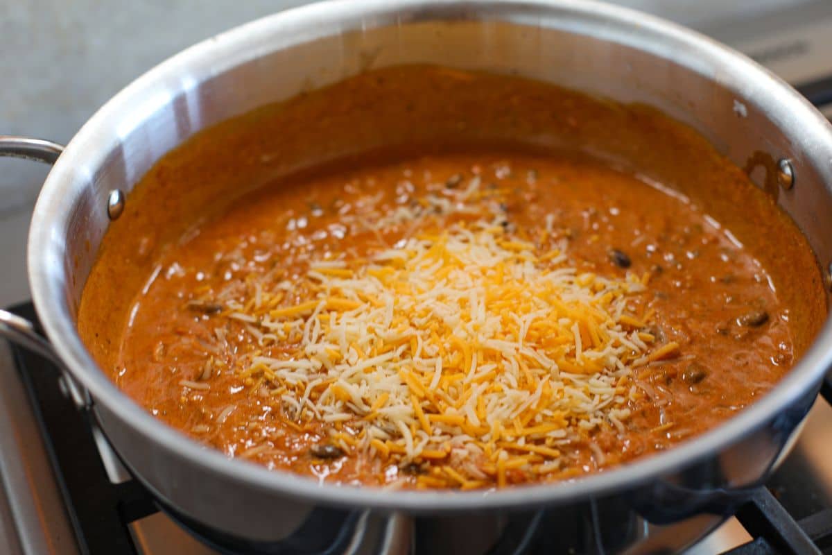A stainless steel pan on a stove contains a creamy, enchilada sauce with black beans, topped with shredded cheese.