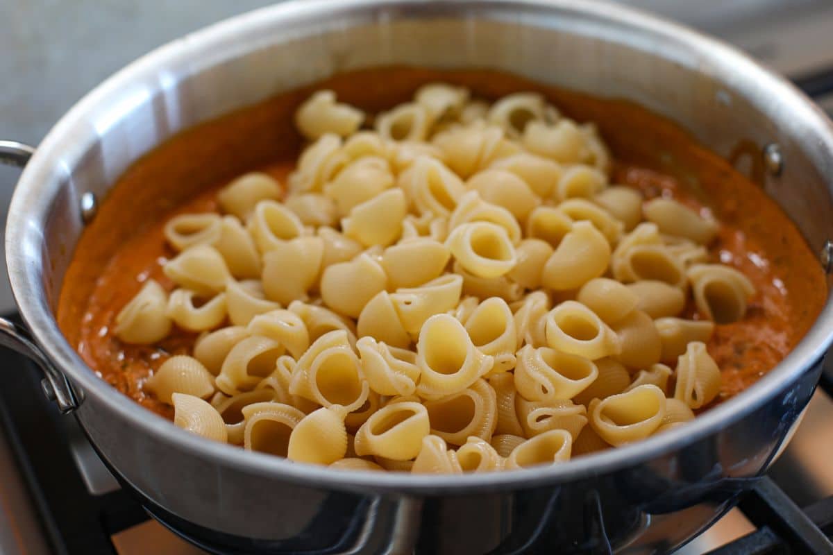 A stainless steel pan on a stovetop contains cooked shell pasta sitting atop a creamy enchilada sauce, creating the perfect base for a delicious Black Bean Enchilada Pasta.