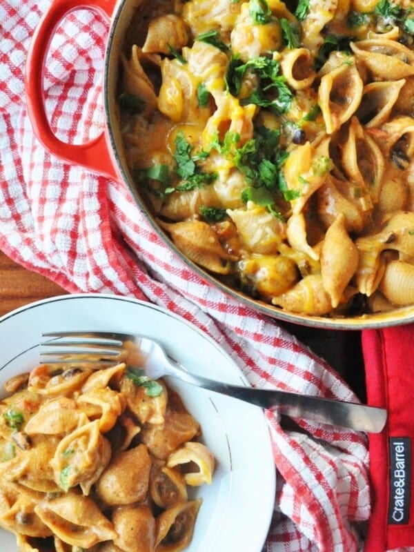 A pot and plate of creamy Black Bean Enchilada Pasta with vegetables, garnished with herbs, placed on a red checkered cloth with a fork on the plate.