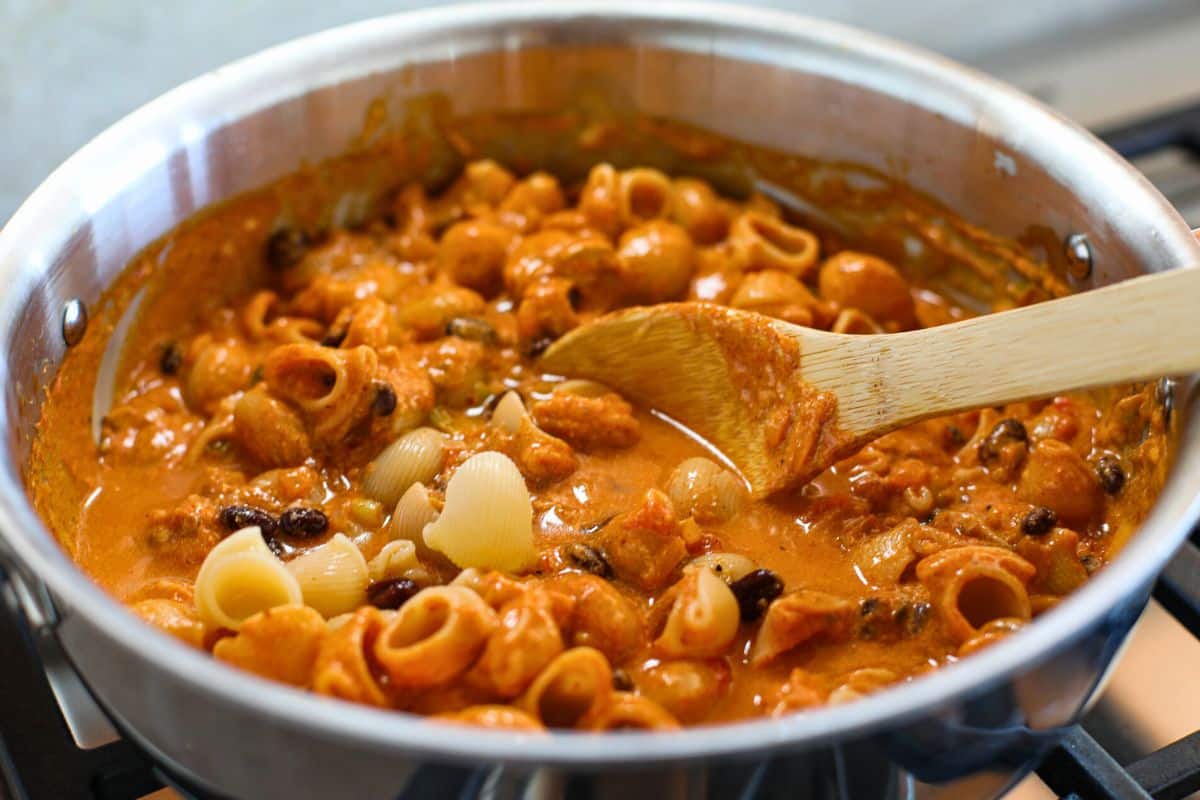 A skillet filled with Black Bean Enchilada Pasta—pasta shells in creamy tomato sauce with beans and vegetables—being stirred with a wooden spoon.