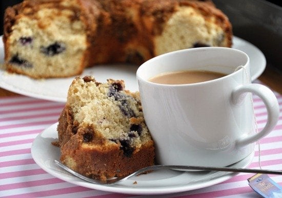 A slice of blueberry cake on a plate with a cup of coffee, set on a pink striped tablecloth.