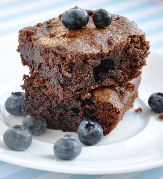 Two chocolate brownies stacked on a plate, topped with and surrounded by fresh blueberries.