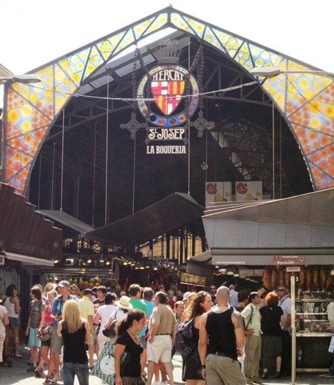 People standing outside Mercat de Sant Josep de la Boqueria, a large indoor market with a colorful stained glass sign above the entrance.