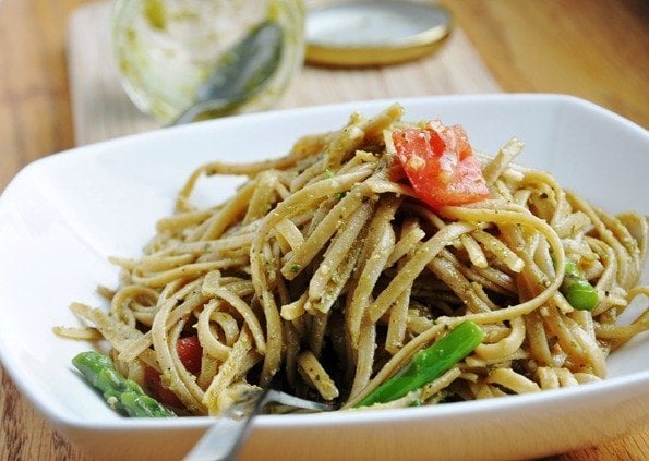 A bowl of pesto pasta with asparagus and tomato pieces. A fork is placed at the front of the dish.