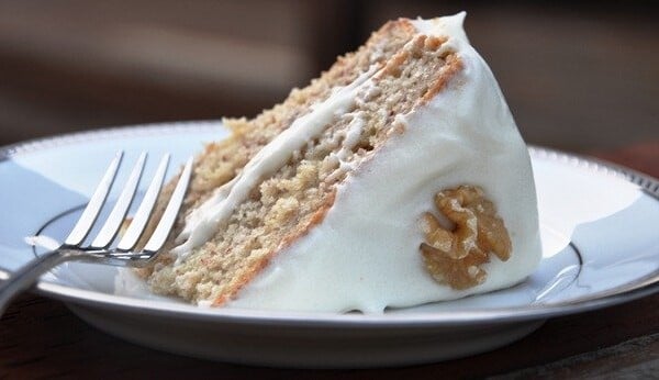 A slice of the worlds best banana cake topped with cream cheese frosting and a walnut sits invitingly on a plate, accompanied by a fork.