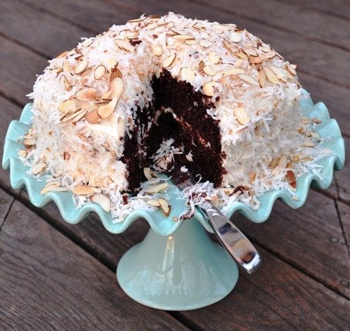 Chocolate cake with frosting, topped with coconut flakes and almond slices, on a light blue cake stand. One slice cut.