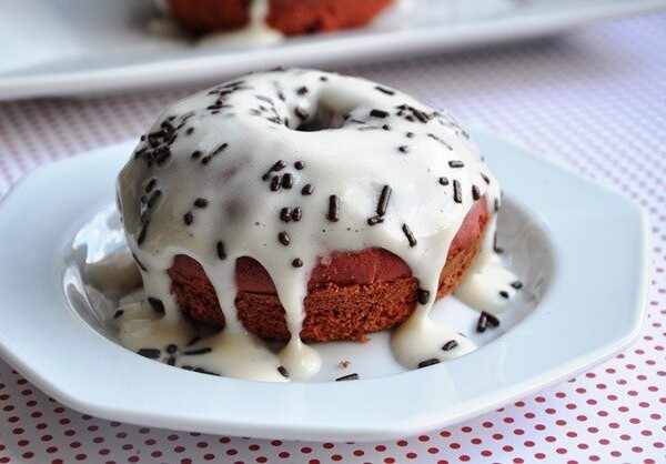 A donut, reminiscent of baked red velvet doughnuts, with white icing and chocolate sprinkles sits enticingly on a white plate atop a polka dot tablecloth.