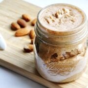 A jar of homemade almond butter sits on a wooden board, surrounded by scattered almonds and accompanied by a white spoon.