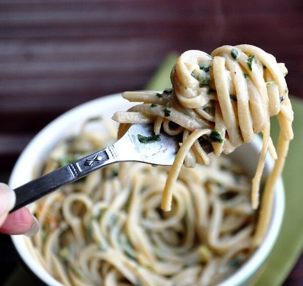 A fork holds a twirled portion of creamy pasta with herbs over a bowl filled with more pasta.