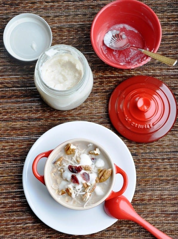 A small red pot containing oatmeal topped with nuts and dried fruits on a white plate, next to a red lid, red spoon, jar of yogurt, and empty red bowl with a fork.