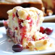 A slice of cranberry orange cake sits on a white plate, topped with a light dusting of powdered sugar. Two whole cranberries are placed nearby on the plate, adding a vibrant touch.