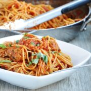 A white bowl of spaghetti with tomato sauce and vegetarian quinoa meatballs, garnished with fresh basil, with a pan of pasta and tongs in the background.