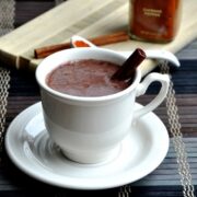 A cup of Mayan hot chocolate in a white teacup with a cinnamon stick, placed on a saucer. A wooden board with cinnamon sticks and a spice container is in the background, enhancing the rich aroma.