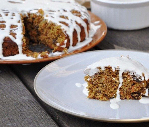 A slice of carrot cake with white icing on a plate. The whole cake is in the background with a piece missing. The cake is on a wooden surface.