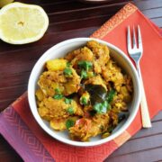A bowl of spiced potato curry garnished with cilantro sits beside Bread Poha on a patterned napkin, with a fork and a halved lemon resting on a wooden surface.
