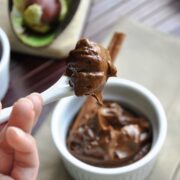 A spoonful of chocolate avocado pudding hovers above a bowl, with an avocado and spoon subtly accentuating the scene in the background.