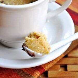A white mug on a saucer holds a 1 Minute Chocolate Chip Cookie in a Mug, resting atop a woven mat. A spoon with the cookie sits invitingly on the saucer.