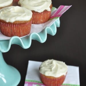 Cupcakes with white frosting displayed on a light blue cake stand and one on a napkin with pink and green stripes.