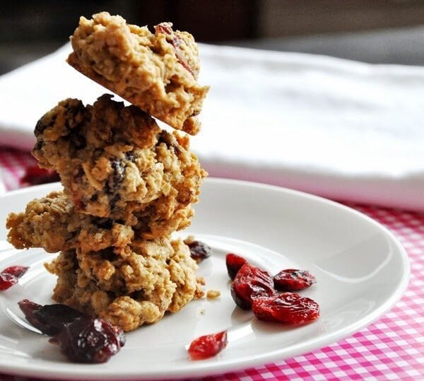 Stacked oatmeal cookies with raisins on a white plate, surrounded by dried cranberries, on a pink checkered tablecloth.