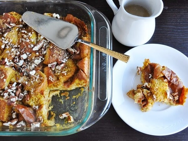 Pumpkin brioche bread pudding in a glass baking dish with a serving spatula, next to a plated portion. A small pitcher of sauce is nearby.