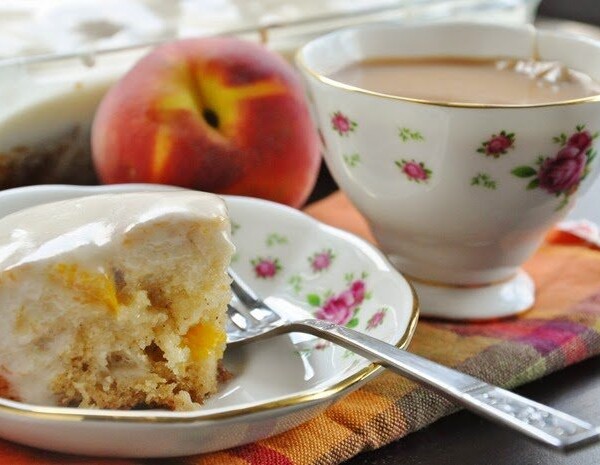 A slice of sweet peach cake with fruit graces a floral saucer, accompanied by a peach and a cup of tea in a matching floral teacup, all set on a plaid cloth.