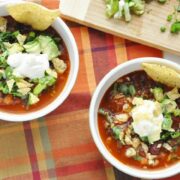 Two bowls of Sweet Potato Bean Chili topped with avocado, sour cream, and tortilla chips rest on a checkered cloth. Nearby, a chopping board holds diced vegetables, adding vibrant colors to the delicious scene.
