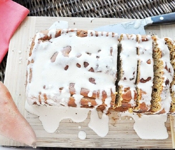 Sliced sweet potato bread with white icing sits on a cutting board next to a knife and a sweet potato, all arranged on a wicker surface.
