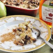 A bowl of apple pie baked oatmeal with pecans and raisins, dusted with cinnamon. A spoon rests in the bowl. Bottles of maple syrup and cinnamon, an apple, and a dish of this cozy oatmeal are in the background.