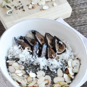 A bowl with sliced figs, shredded coconut, chia seeds, and almond slices is complemented by cardamom pods on a cutting board nearby—an enticing blend reminiscent of cardamom rosewater oatmeal that infuses your morning with aromatic spice.