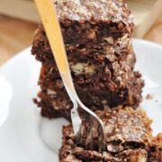 A fork pierces a stack of three flourless chocolate walnut brownies, each rich with nuts, resting on a white plate. The background reveals foil and a wooden board.