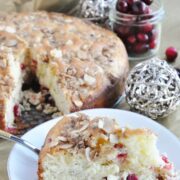 Slice of cranberry almond coffee cake on a white plate, with the rest of the cake behind it and a jar of cranberries nearby. Silver decorative spheres add a touch of elegance to this delightful scene.