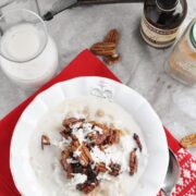 A bowl of pecan pie coconut oatmeal adorned with crunchy pecans and coconut flakes sits beside a glass of milk, a bottle of vanilla extract, and syrup on a red cloth, with decorative silver ornaments shimmering in the background.