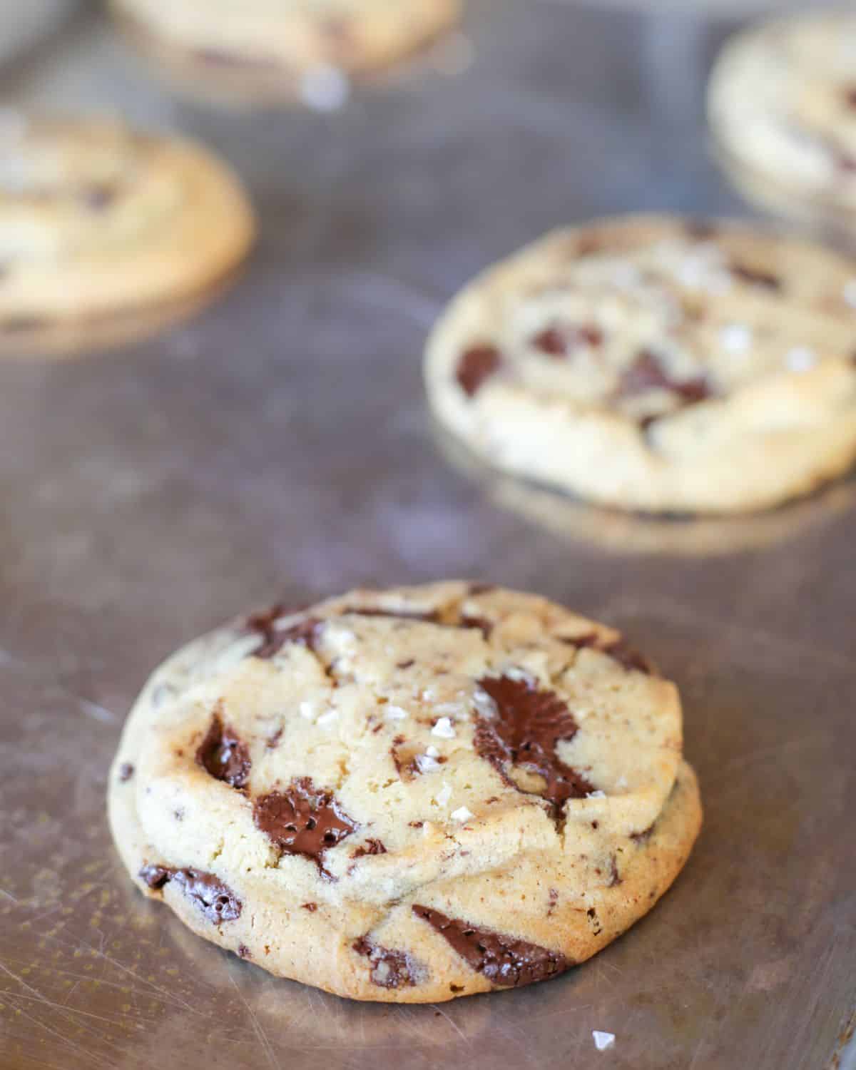 A close-up of The New York Times Chocolate Chip Cookies with visible chocolate chunks and a sprinkle of sea salt on a metal baking sheet.