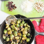 A skillet with cranberry almond roasted Brussels sprouts sits on the stovetop. Nearby, shaved almonds, grated cheese, and chopped cranberries rest on a green cutting board, while a red oven mitt adds a pop of color.