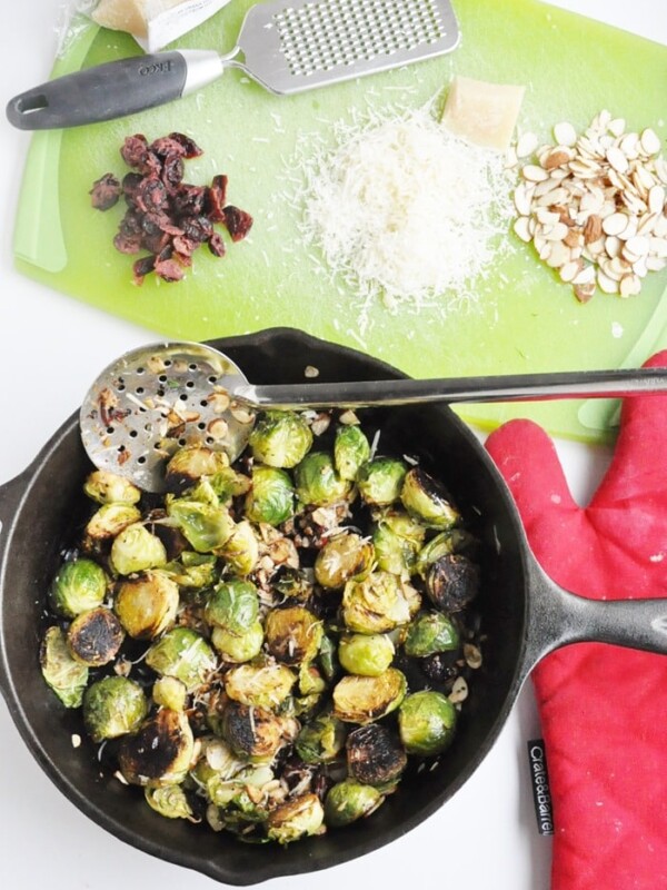 A skillet with cranberry almond roasted Brussels sprouts sits on the stovetop. Nearby, shaved almonds, grated cheese, and chopped cranberries rest on a green cutting board, while a red oven mitt adds a pop of color.