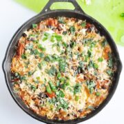 A skillet with a baked casserole topped with herbs and cheese, featuring Black Bean Mexican Skillet Eggs. Nearby, chopped avocado, cilantro, and grated cheese rest on a green cutting board.