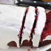 Red velvet banana bread loaf with white icing, partially sliced, on a white plate. A knife is placed beside this mouthwatering creation.