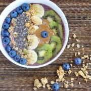 A blueberry banana acai bowl topped with kiwi slices, chia seeds, granola, and almond butter rests on a textured surface, scattered with extra granola and fresh blueberries.