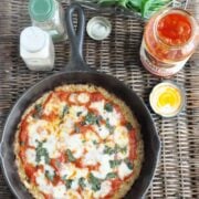 A cast iron skillet holds a delicious homemade crispy cauliflower pizza topped with cheese and basil, surrounded by a jar of pizza sauce and various seasonings on a wicker surface.