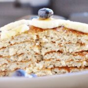 A stack of four high protein banana bread pancakes topped with banana slices, a blueberry, and syrup rests on a plate, accompanied by a few scattered blueberries.